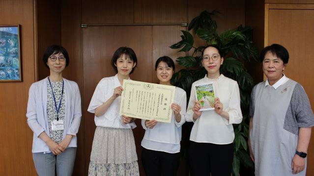 表彰状と作成した冊子を手にもつ学生、豊沢准教授、岡本学長が立って並んでいる